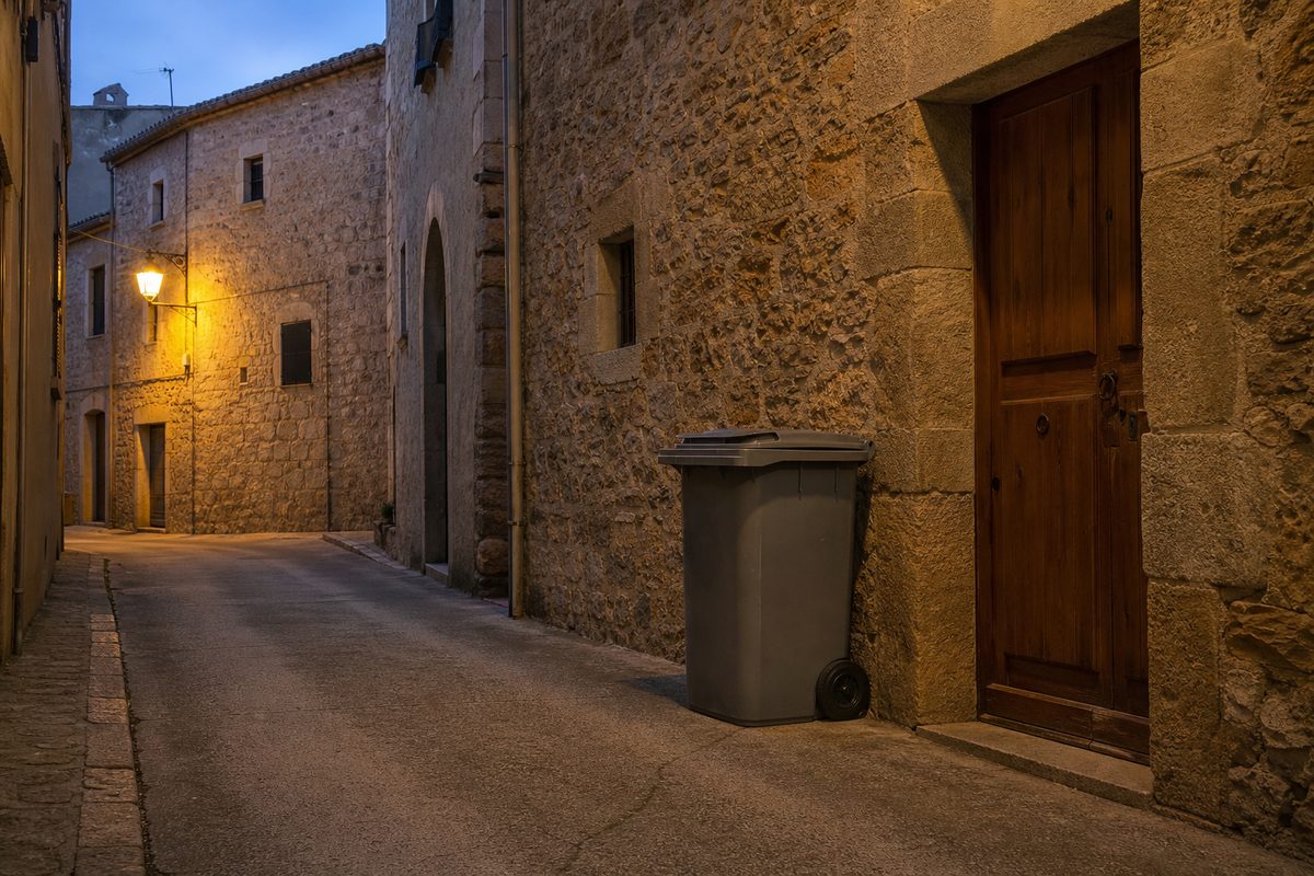 Cubo de residuos gris junto a la puerta de una casa de piedra en una calle estrecha al anochecer